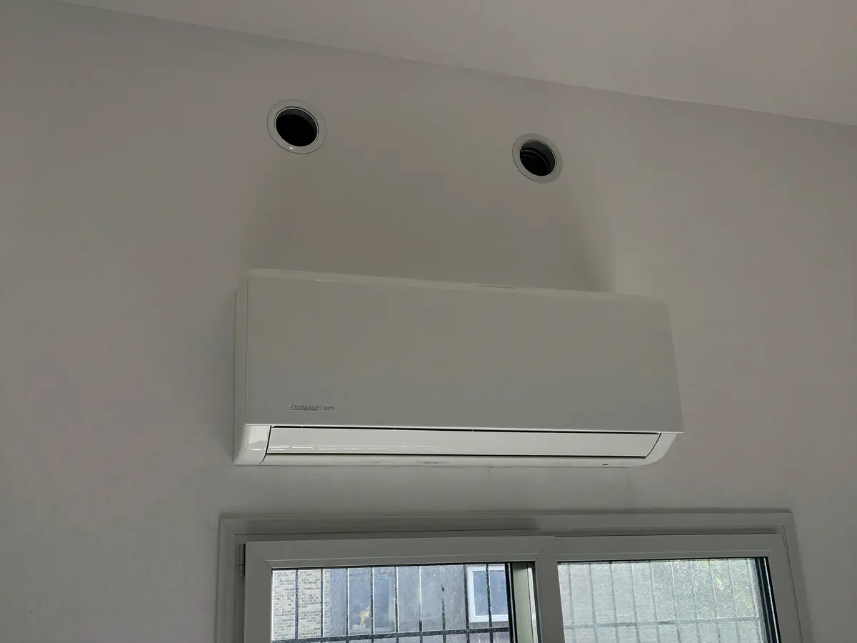 Vaste airco zonder buitenunit onder een raam met twee ronde roosters in de muur