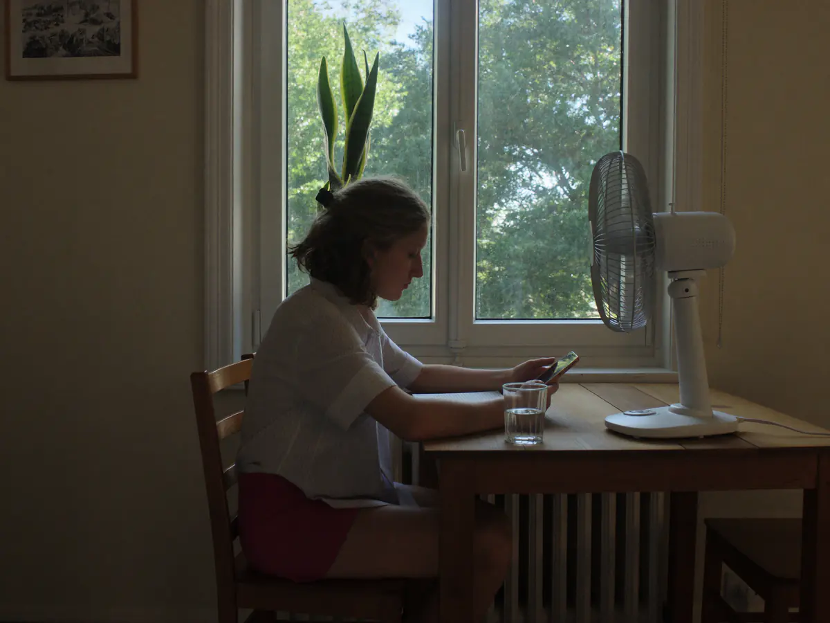 Vrouw aan de keukentafel met haar telefoon, een glas water en een tafelventilator op een warme dag
